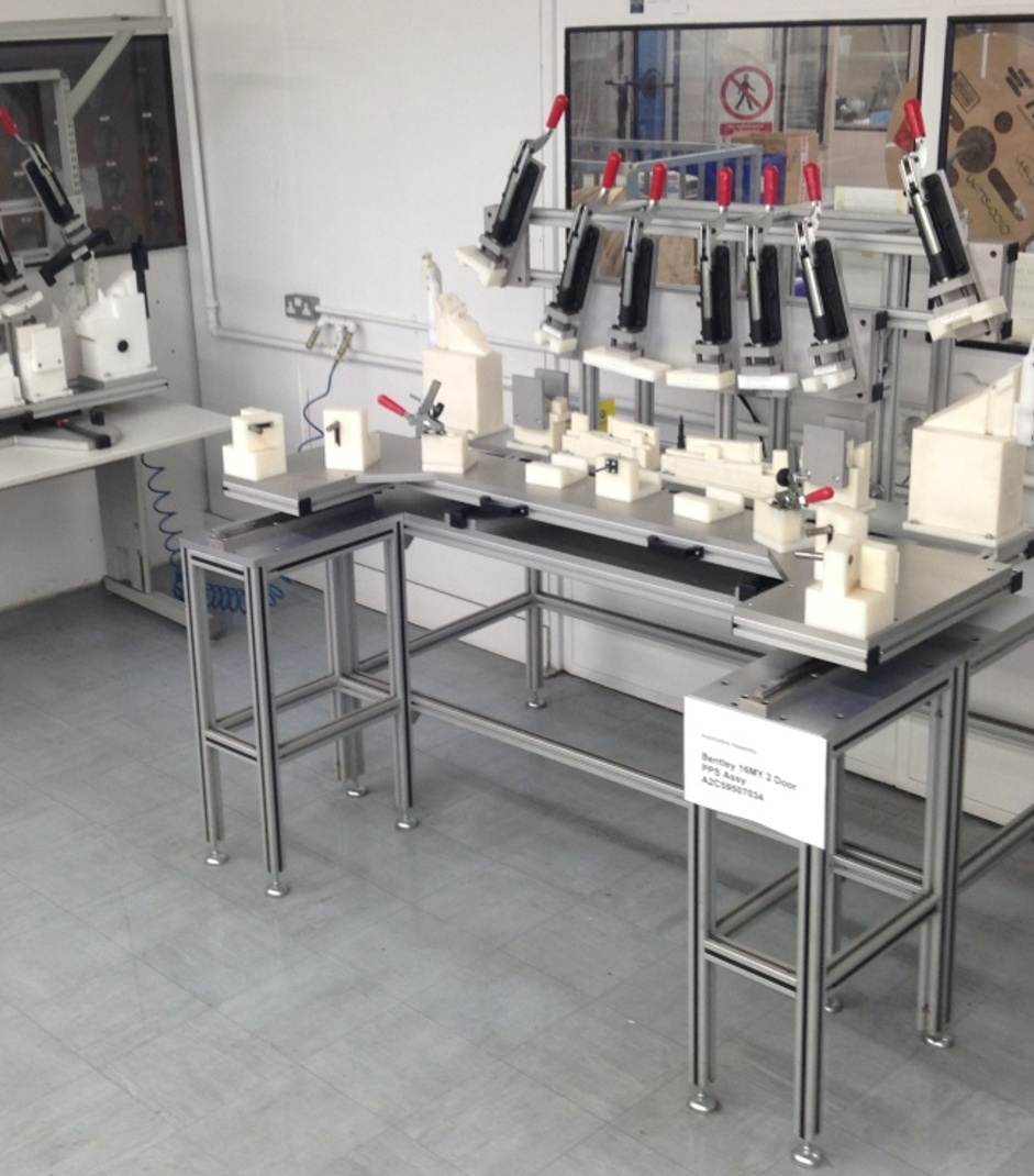 A workstation with multiple mechanical clamps and fixtures mounted on a metal frame table in a clean, industrial environment. There is a white sign on the table leg, and various tools and equipment are organized neatly on and around the table.